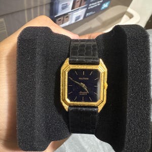 May include: A Waltham wristwatch featuring a gold-tone octagonal case with a dark blue dial, gold hands, and gold hour markers. The watch face displays the text Waltham and Maxine Quartz. It is fitted with a black textured leather strap.
