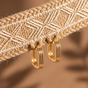 Peut inclure: Une paire de créoles dorées de forme rectangulaire avec une texture à trois nervures. Les boucles d'oreilles sont suspendues à une bande de tissu tissé beige présentant un motif géométrique brodé blanc sur toute sa longueur.