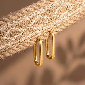 May include: Two gold-toned hoop earrings with an elongated, rectangular shape. The earrings are suspended from a textured, woven beige fabric strap that features a subtle, repeating geometric pattern in a lighter shade of tan.