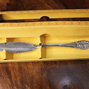 Può includere: Un coltello da burro argentato con una lama a forma di foglia e un manico ornato è contenuto in una scatola di cartone gialla. La scatola ha una finestra rettangolare e un bordo arancione. Il coltello è esposto su un interno giallo.