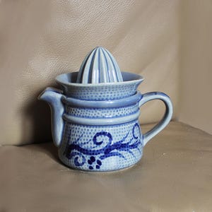 May include: A blue and white ceramic citrus juicer. The juicer has a conical reamer and a small spout. The body of the juicer is decorated with a blue floral design. The juicer has a handle and is sitting on a neutral surface.