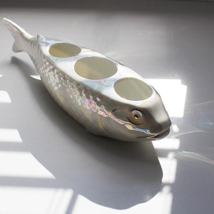 Pode incluir: Uma peça decorativa em cerâmica perolada em forma de peixe com três aberturas circulares. O peixe tem um padrão de escamas detalhado e um olho dourado. O objeto está sobre uma superfície branca, com sombras.