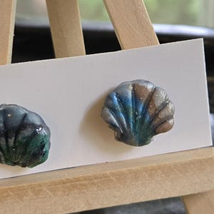 May include: A pair of stud earrings shaped like seashells, each featuring a gradient of metallic blue, green, and silver tones. The earrings are attached to a rectangular white display card, which is positioned on a small wooden easel.