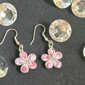 May include: A pair of silver-toned drop earrings featuring a five-petal flower design with pink and white marbled enamel inlay, suspended from silver-toned ear wires, surrounded by several clear, faceted, round decorative glass gems.