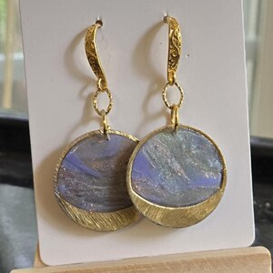 May include: A pair of dangle earrings featuring circular pendants with a marbled purple, blue, and silver finish. Each pendant is framed by a textured gold-tone metal crescent at the base and hangs from an ornate, engraved gold-tone hook.