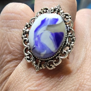 May include: An oval ring featuring a smooth, polished cabochon with a swirling pattern of deep purple and white colors. The stone is set within an intricate, silver-toned metal frame that displays detailed scrollwork and filigree patterns.