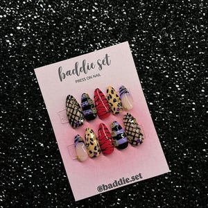 Puede incluir: Un juego de diez uñas postizas en forma de almendra sobre una tarjeta rosa claro con la etiqueta baddie set PRESS ON NAIL y @baddie.set. Las uñas presentan diseños variados que incluyen red negra, rayas moradas y negras, estampado de tigre rojo y leopardo.