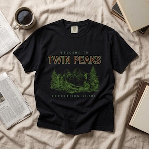 Könnte beinhalten: Ein schwarzes Kurzarm-T-Shirt mit einer grünen Grafik von Bergen und Tannenbäumen. Der Text "WELCOME TO TWIN PEAKS" steht in fetter Schrift über der Landschaft und "POPULATION 51,201" in kleinerer Schrift unter der Waldillustration.