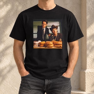 May include: A black short-sleeved t-shirt featuring a rectangular graphic print of two men in suits, one wearing a cowboy hat, standing behind a table filled with various glazed and powdered donuts. The shirt has a crew neckline and a standard fit.