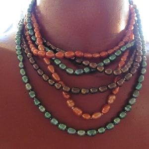 Könnte beinhalten: Eine mehrreihige Halskette aus grünen und orangefarbenen ovalen Perlen. Die Kette ist vielschichtig und hat einen lässigen, bohemian Stil.