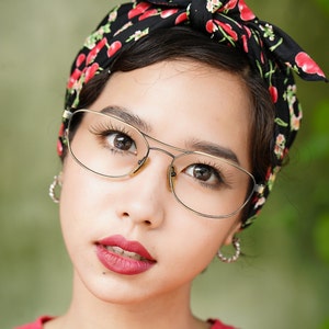 Op de afbeelding: Een vrouw met donker haar draagt een zwart hoofdband met rode kersen erop gedrukt. Ze draagt een gouden bril en heeft rode lippenstift op. Ze draagt een rood shirt.