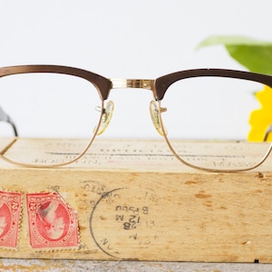 May include: Vintage brown and gold eyeglasses with a butterfly design on the arms. The glasses are resting on a wooden box with stamps.