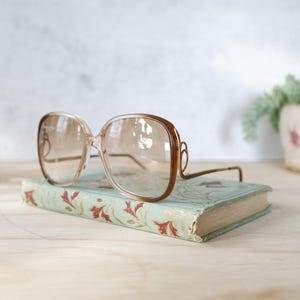 Puede incluir: Gafas de sol cuadradas de gran tamaño con un tinte marrón claro sobre un libro antiguo. Las gafas tienen montura y patillas marrones. El libro es de color verde claro con un estampado floral y un aspecto desgastado. Una pequeña planta en maceta está en el fondo.