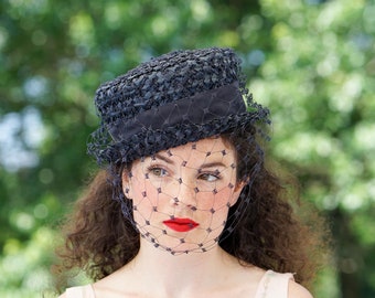 May include: A woman with curly dark hair wears a black hat with a veil. The hat is a pillbox style with a wide brim and a black ribbon around the crown. The veil is made of fine netting and covers her face.