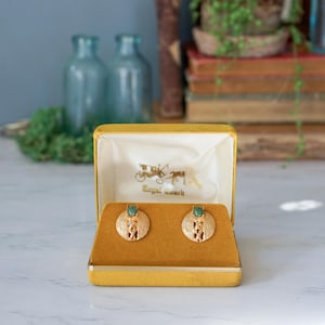 Puede incluir: Un par de pendientes de tono dorado con detalles de piedras verdes en una caja de terciopelo amarillo. La caja tiene el texto "Royal Coach" impreso en el interior.