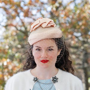 May include: A vintage-style hat with a peach-colored bow and a delicate veil. The hat is a light beige color and has small polka dots. The person is wearing a white cardigan and a light blue dress. The background is a blurred view of trees.