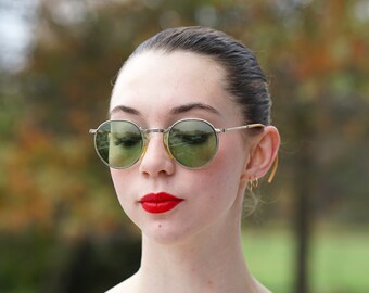 May include: Round, silver-framed sunglasses with green lenses. The sunglasses are worn by a person with red lipstick and small gold hoop earrings. The background is blurred with green and brown tones.