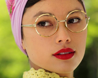 Könnte beinhalten: Eine Frau mit dunklen Haaren trägt ein rosa-weißes Blumenkopftuch und eine runde goldene Brille. Sie trägt einen gelben Häkelpullover.