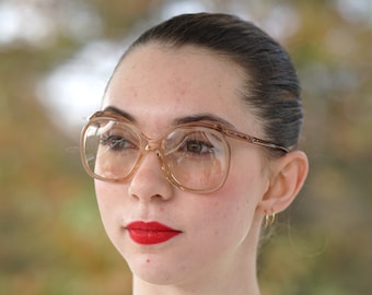 May include: A woman with dark hair wearing large, brown-rimmed glasses and red lipstick. She is wearing a white, linen-like shirt.