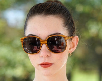 Könnte beinhalten: Quadratische Sonnenbrille mit gestreiften Holzmaserungsrahmen. Die dunklen Gläser spiegeln die Umgebung wider. Die Person trägt ein cremefarbenes Oberteil mit braunem Kragen und Schleife.