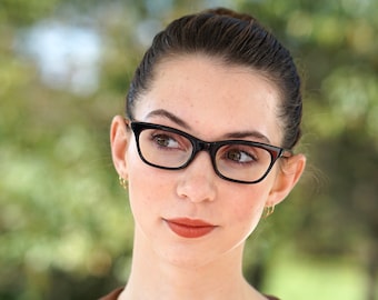 May include: A young woman with brown hair pulled back in a bun, wearing black cat-eye glasses, a brown collar, and a beige top with a gold bow.