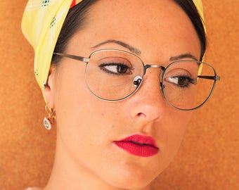 Peut inclure: Une femme aux cheveux foncés porte un foulard jaune à imprimé floral rouge et vert noué sur le dessus de sa tête. Elle porte des lunettes à monture argentée et a du rouge à lèvres rouge. Elle porte un pull rose.