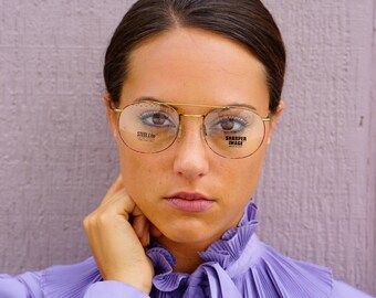 May include: A woman wearing a purple blouse with a ruffled collar and gold-rimmed round glasses. The glasses have the text "STEEL-LITE" and "SHARPER IMAGE" printed on the lenses.