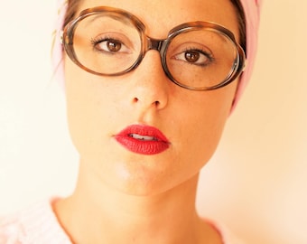 May include: A woman with brown hair is wearing a pink headscarf with floral embroidery and large, round, brown-rimmed glasses. She is wearing a pink sweater and has red lipstick on.