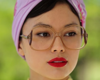 May include: A woman wearing a purple turban with a floral design and large brown framed glasses. She is wearing a yellow crocheted sweater.