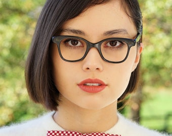May include: A woman with short black hair wearing black framed glasses and a white sweater with a red and white checkered bow tie.