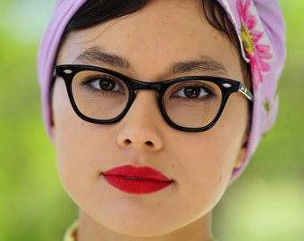 May include: A woman with dark hair is wearing a light purple headscarf with a floral print and black cat-eye glasses. She is wearing a yellow crocheted sweater.