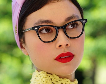 May include: A woman wearing a light purple headband and black cat-eye glasses. She is wearing a yellow crocheted sweater.