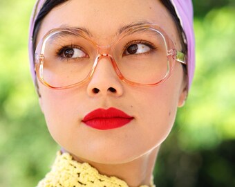 May include: A woman wearing a purple headband with a floral pattern and large, clear, plastic framed glasses. She is wearing a yellow crocheted sweater and has red lipstick.