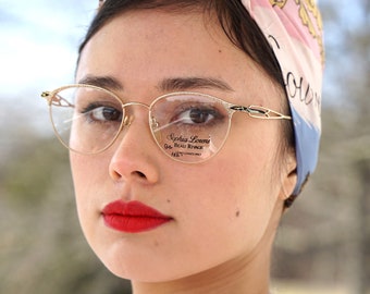 Peut inclure: Une femme portant une chemise rose et un foulard coloré à motifs floraux. Elle porte des lunettes à monture dorée avec le texte "Sophia Loren's 94 - Beau Rivage 145 - Lenses Only" visible dans le reflet.