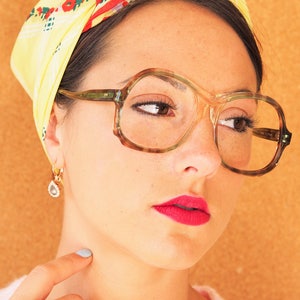 Puede incluir: Una mujer con lápiz labial rojo lleva gafas grandes, redondas, de color verde y un pañuelo amarillo con estampado floral rojo y verde.
