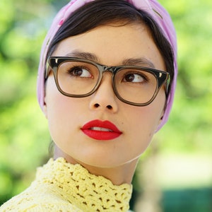 May include: A woman with dark hair wearing a purple headscarf and black framed glasses. She is wearing a yellow crocheted sweater and has red lipstick on.