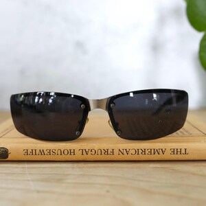 May include: Black sunglasses with a silver bridge and dark lenses rest on a vintage book titled "THE AMERICAN FRUGAL HOUSEWIFE". The sunglasses have a rectangular shape and are secured with small rivets.