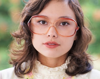 May include: A woman with brown hair wearing large, brown, oval-shaped glasses and a white dress with floral embroidery.