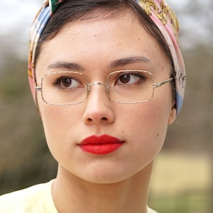 May include: A woman with dark hair wearing a pink, blue, and gold patterned headscarf and square-shaped glasses with a silver frame. She is wearing red lipstick and a light yellow shirt.