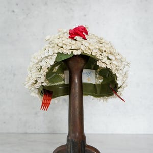 Puede incluir: Un sombrero floral blanco vintage con un acento rojo, exhibido en un soporte de sombrero marrón. El sombrero presenta un interior verde y un pequeño peine rojo. El soporte del sombrero tiene una base de trípode.