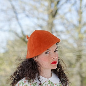 May include: A woman with curly brown hair is wearing a bright orange felt hat with a pointed tip. She is looking to the side and has red lipstick on.