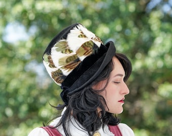 May include: A woman wearing a black velvet top hat with a white and green feather trim. The hat is tilted slightly to the side. She has long, dark hair and is wearing a white blouse with a ruffled collar and a burgundy vest.