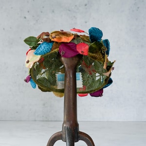 May include: A vintage hat with a green mesh base, adorned with colorful fabric flowers in shades of red, blue, orange, and purple. The hat is displayed on a dark brown, three-legged hat stand against a neutral background.