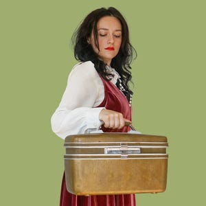 May include: A vintage, rectangular, bronze-colored suitcase with white trim and a silver clasp. The suitcase is held by a person wearing a red velvet dress and a white blouse. The background is a solid, light green color.
