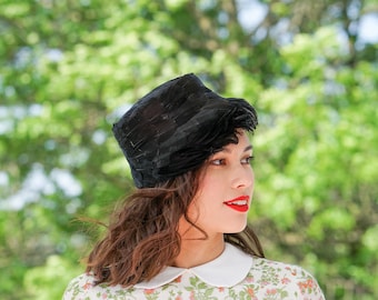 May include: A woman with dark hair wearing a black hat with a feather trim and a white collared dress with a floral pattern. She is looking to the right and has red lipstick on.
