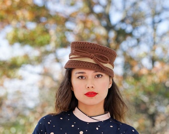 May include: A woman wearing a brown knitted hat with a beige ribbon and bow. She is wearing a navy blue dress with white polka dots and has red lipstick.