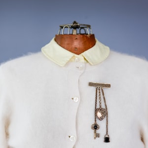 Puede incluir: Un cárdigan blanco roto con cuello amarillo claro, abotonado. Un broche decorativo con colgantes, incluyendo un corazón, una llave y un bolso, está prendido en la parte delantera. El broche es de metal dorado.