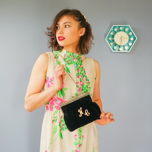 May include: A woman wearing a floral dress with a black velvet clutch bag with gold hardware and the initials "H.E." embroidered on the front.