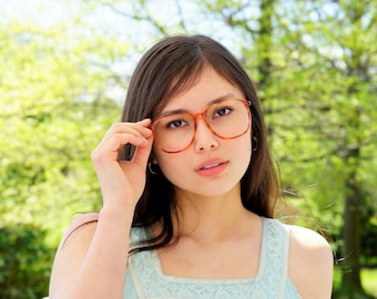 Puede incluir: Una joven con el pelo largo y castaño que lleva un vestido azul claro sin mangas y unas gafas grandes, redondas y de carey. Está mirando a la cámara con una expresión seria.