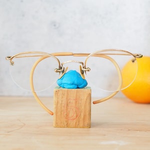 Peut inclure: Lunettes sans monture dorées avec un design incurvé unique, reposant sur un bloc de bois. Les lunettes ont des verres transparents et sont maintenues ensemble par une fine barre métallique.
